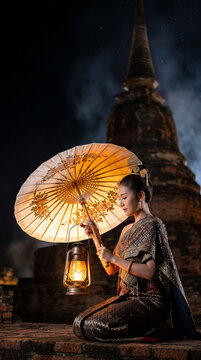 Beautiful Thai woman in traditional dress holding paper umbrella and oil lamp at ancient temple ruins.