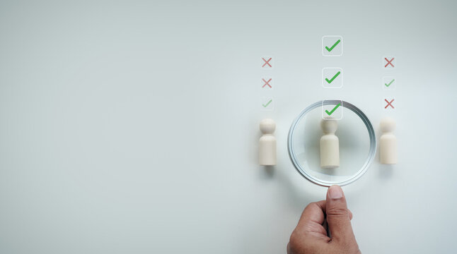 Hand holding magnifying glass focusing on best wooden peg doll candidate with green check mark icons overlay among figures on white background, concept of HR recruitment and selection.