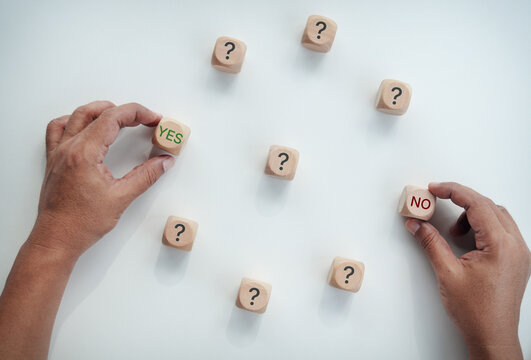 Hands holding wooden blocks with YES and NO text among question mark blocks on white desk, concept of business decision making, choice selection, problem solving, and direction.