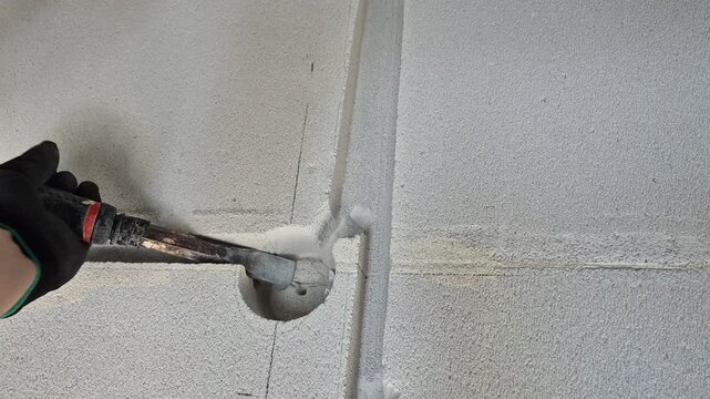Close-up of a construction worker hands wearing protective gloves chiseling a groove in a gas concrete wall with a hammer.