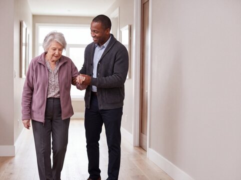 Male caregiver assisting senior woman walking down home hallway