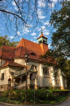 A side view of the church at Babinski Clinical Hospital in Krakow, Poland. A steep red tiled roof and green-patinated bell tower rise above pale walls, framed by branches against a blue sky.