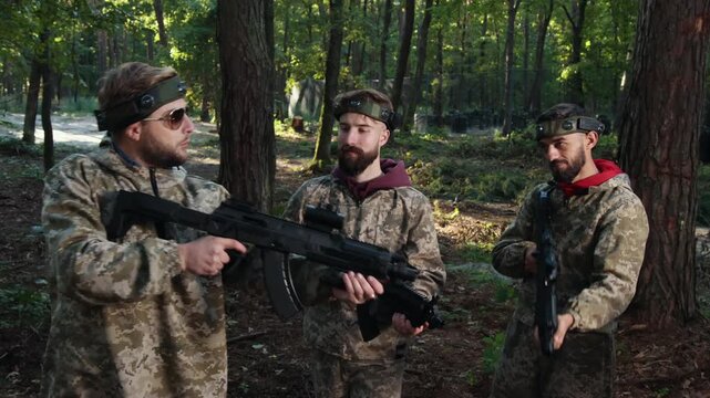 Young people, Camouflage gear, Tactical gear. Cluster Laser tag players, clad in camouflage gear and each wielding marker, assembles in forest environment, intently engaged