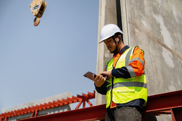Construction worker taps a tablet beside a precast concrete panel beneath an overhead crane hook, using mobile technology at a construction yard © KL 1981