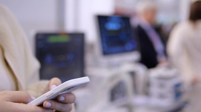 Person holds smartphone in focus. Background shows blurred monitors and people. Workplace or tech environment implied. Soft focus emphasizes mobile use over surroundings