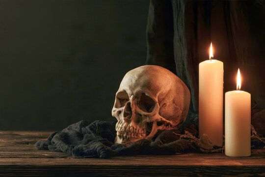 moody gothic still life photography featuring a human skull resting next to glowing white candles and dark draped fabric on a rustic wooden table against a dark background
