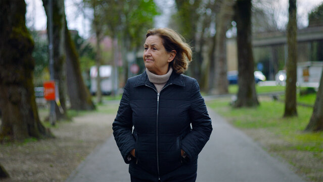 Caucasian senior woman walking on a tree-lined park path with hands in pockets, peaceful stroll and wellbeing