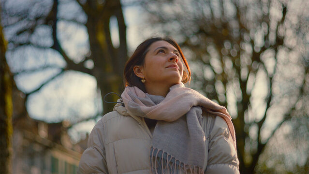 Serene Caucasian woman looking up at bare trees at golden hour, peace, gratitude and connection to nature