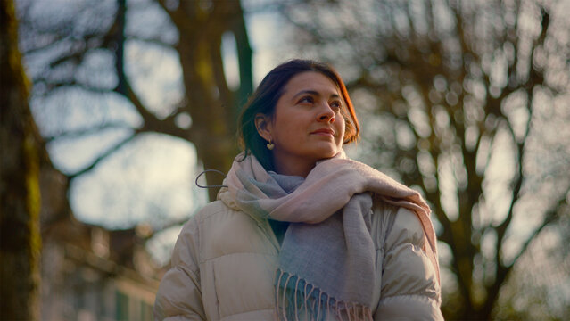 Serene Caucasian woman looking up at bare trees at golden hour, peace, gratitude and connection to nature