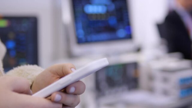 Person holds white smartphone. Fingers tap screen. Background shows blurred monitors. Office or tech workspace setting