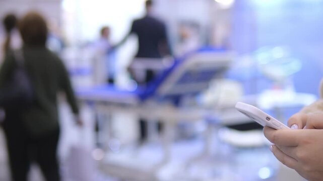 Person holds smartphone, focused on screen. Background shows blurred medical setting with staff and equipment. Ambient lighting reflects clinical environment. Blurred figures move around patient bed