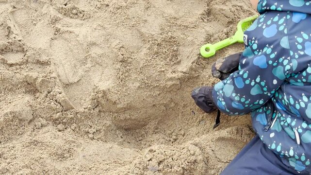 A small child digs happily in fine sand. Gloved hands scoop dirt with focused joy near a plastic shovel. The large sandbox sits in a sunny park.