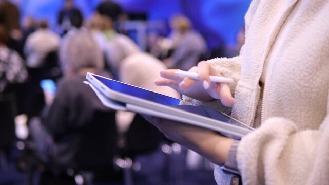 Woman holds tablet and stylus. She writes notes in event setting. Background shows blurry attendees. Focus on her hands and device. Scene suggests professional or educational environment