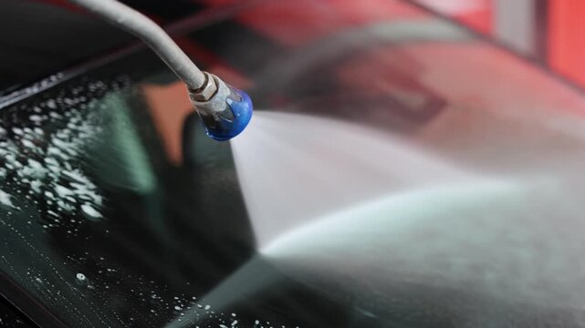 Pressure washer cleans the windshield of a passenger car at a self service car wash. Water spray removes dirt from the front glass surface. Vehicle washing process covers an important visibility area.