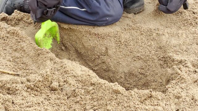 A child digs playfully with a green shovel. Hands move sand with focused delight. The deep trench sits in a sunny sandbox.