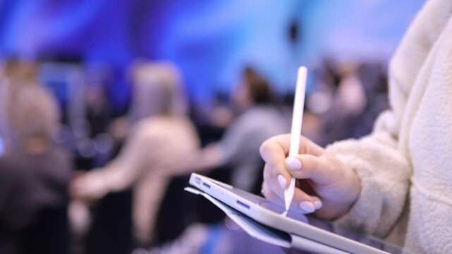 Person uses stylus on tablet during presentation. Background shows blurred audience in dim blue-lit room. Focus on hand and device with soft lighting