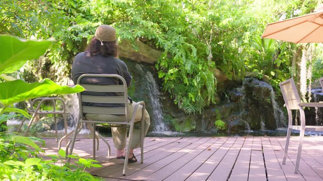 A person from behind relaxes on a patio chair, finding solitude and tranquility while contemplating a beautiful waterfall in a lush, landscaped garden oasis.