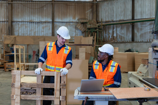 Wood workshop: woodworking carpenter discusses plywood with his male colleague beside a laptop and a wooden pallet.