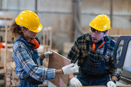 Wood workshop: woodworking carpenter and colleague examine plywood together as the woman holds a piece beside the panel saw.