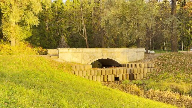 A stone bridge spans a small stream flowing through grassy autumn slopes. A quiet view in Catherine Park, Pushkin, Russia inspires peaceful calm and warm nostalgia.