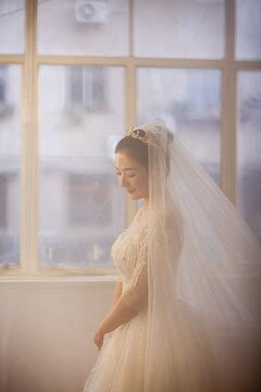 Beautiful Asian bride in white beaded wedding gown with tiara and veil, soft window light, wedding preparation portrait