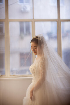 Beautiful Asian bride in white beaded wedding gown with tiara and veil, soft window light, wedding preparation portrait