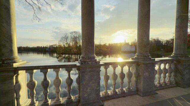 Marble columns and a balustrade frame a serene view of the Turkish Bath pavilion at golden sunset. A peaceful scene in Catherine Park, Pushkin, Russia brings warm tranquil joy.