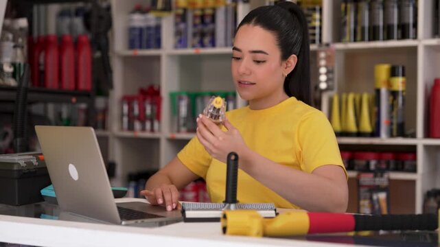 Young latina female mechanic checking auto parts and typing on a laptop inside a car repair shop