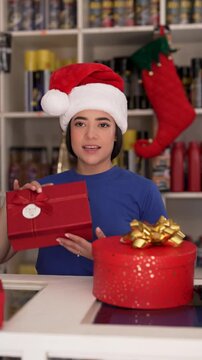 Cheerful latina retail worker wearing santa hat packing and preparing red holiday gifts in store