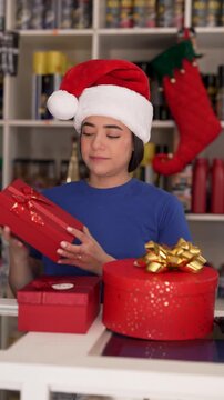 Young latina saleswoman wearing santa hat organizing festive red present boxes in gift shop
