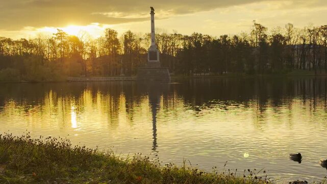 The tall Chesme Column stands gracefully in the middle of a sunlit lake. A peaceful slow motion view in Catherine Park, Pushkin, Russia inspires quiet awe and warmth.