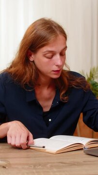 Young woman on home table reads book and marks key line during calm study at home. Adult lady on home table learns with laptop and notes staying focused on lesson for skill growth and exam prep daily.