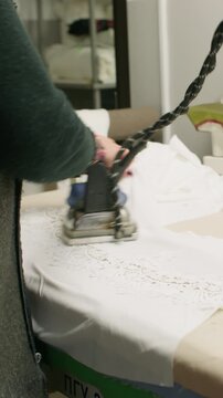 Vertical video. Dressmaking process. Ironer smoothing textile. Woman seamstress steaming white lace fabric with professional appliance in tailor workplace.
