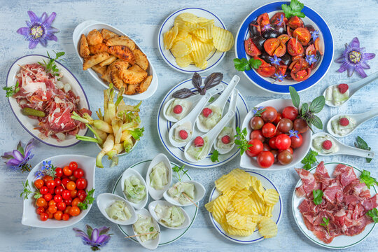 Different plates of food are arranged on a table. There are appetizers like olives, meats, and fresh vegetables. Some plates are filled with chips and small salads. Flowers decorate the scene.