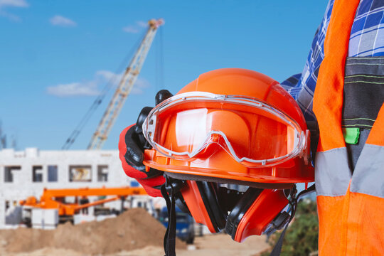 Construction worker holding orange safety helmet with goggles and ear protection at building site. Industrial PPE, occupational safety or construction equipment concept.
