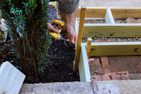 Builder uses drill to secure wooden steps pieces together near shrubs
