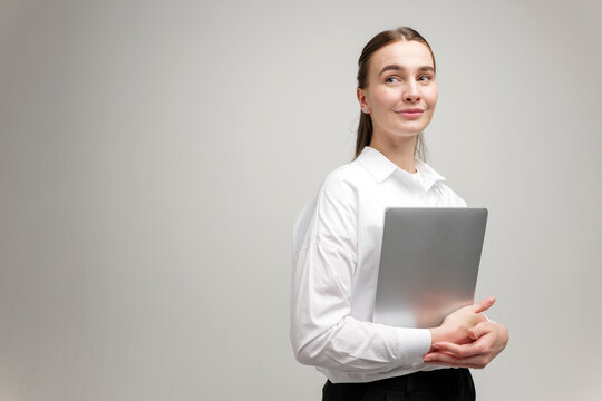 Young woman in formal attire confidently holds laptop while looking sideways in a bright modern office