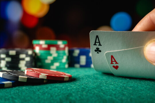 Poker player holds ace of clubs and ace of hearts over green table felt, with chips nearby. Strong starting hand creates anticipation in casino card game under colorful blurred lights.