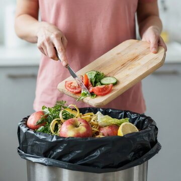 Person scraping fresh vegetable scraps from a wooden cutting board into a kitchen trash can filled with food waste
