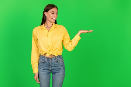 Smiling Young Woman In Yellow Shirt Pointing At Open Hand With Joyful Expression, Standing In Front Of Green Background, Perfect For Presentations Or Advertisements. Business Concept.