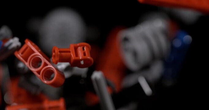 Slow motion Orange construction toy parts float against black, with gray pieces behind them. Building components rotate slowly, revealing holes and connector shapes in dramatic close up.
