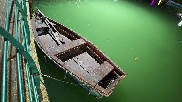 Old wooden rowboat moored on calm green water of a city park pond at nighttime, tied with a chain beside a green metal railing and stone embankment, romantic quiet evening leisure and relaxing boating