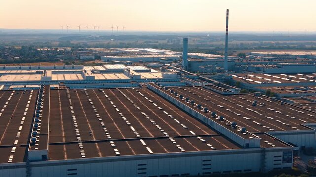 Factory rooftops at sunset. An aerial view of a factory complex with long rooftops and smokestacks at sunset, with wind turbines on the horizon.