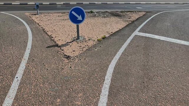 Blue round keep right traffic sign with a bold white arrow on a sandy triangular island between two diverging white lines on dark gray asphalt at a road fork, with a roadway and curb in the background