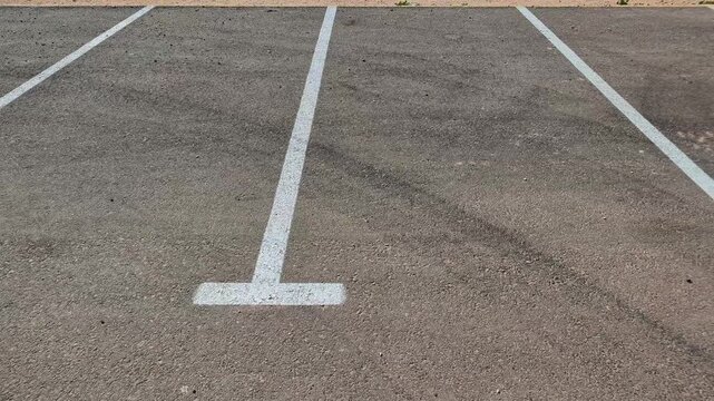Empty parking spaces on gray asphalt marked with white painted lines, one line ending in a T-shaped bar, vacant parking spots in an outdoor car park, free public parking lot with clear road markings
