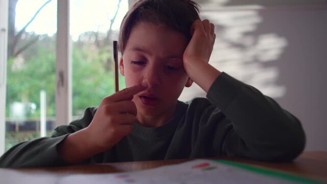 Frustrated Caucasian schoolboy leans his head on his hand and taps his pencil while doing homework at the table, tired of studying, learning difficulties.