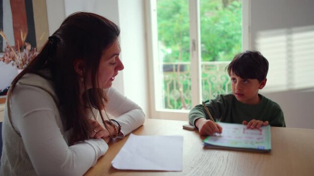 Caucasian mother sits opposite her son and supervises his homework, pointing at his workbook and explaining, parental involvement in education.