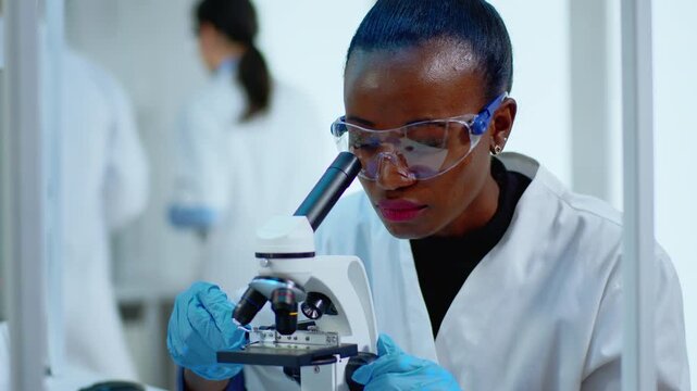 Microscope, scientist and analysis as a woman researcher leans toward the instrument in safety glasses and gloves; scientific observation, specimen testing and biomedical discovery.