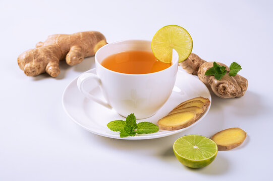White cup of herbal ginger tea garnished with lime and mint, surrounded by fresh ginger rhizomes and slices. Healthy wellness beverage isolated on white background.