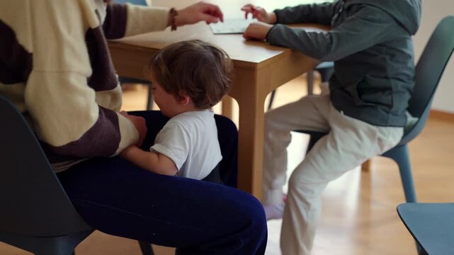 Caucasian mother holds her baby at the table while supervising and guiding her son's homework, mom as teacher and supervisor of her children's education.